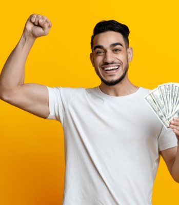 Saving money, gambling, gaming, loan concept. Happy middle-eastern guy in white t-shirt holding bunch of money, raising fist up and smiling, won lottery, yellow studio background, copy space